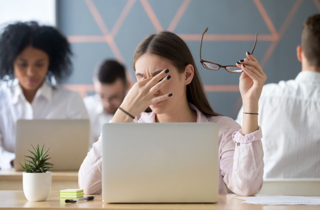 A young person removing their glasses and rubbing their eyes while in front of their laptop screen in a classroom setting.