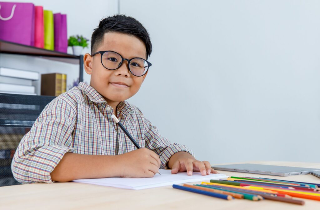A school-age child sitting a desk colouring while wearing glasses.