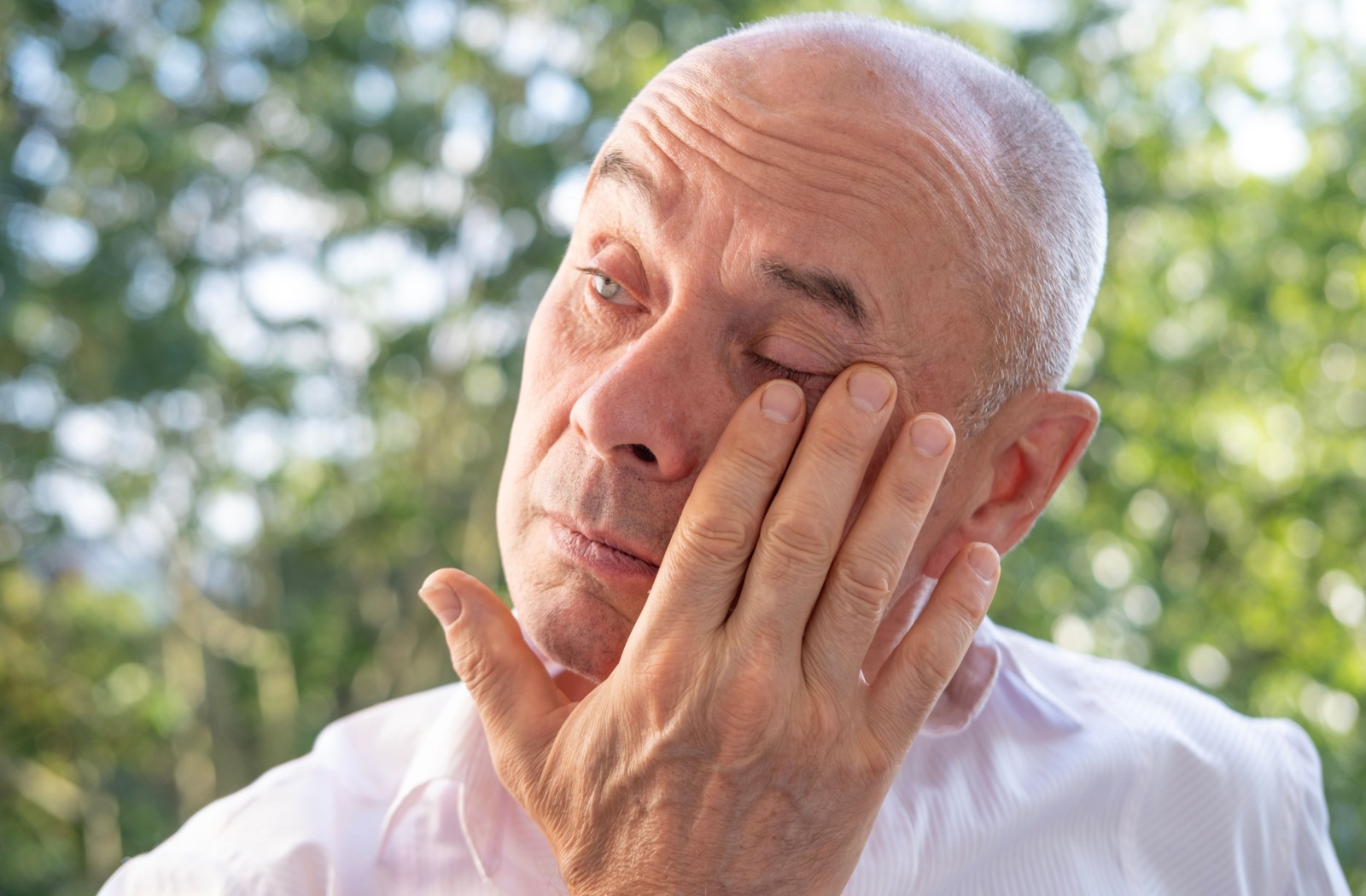 A person rubs their eye in discomfort while standing outdoors, likely from allergies or dry eye.