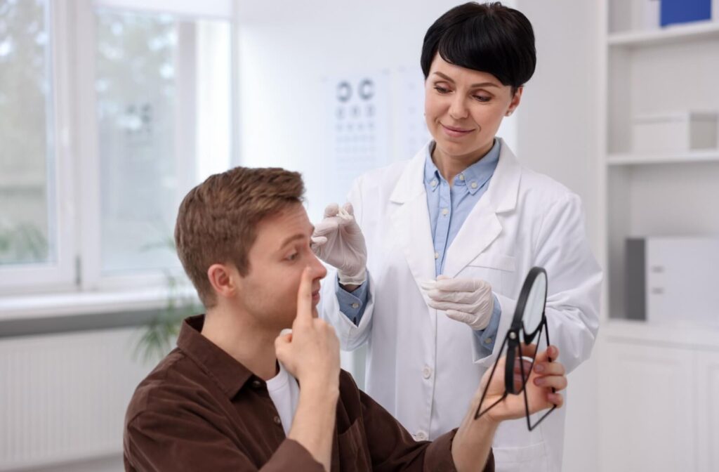 Optometrist assisting a patient in a clinic as he practices inserting a contact lens while holding a small mirror, demonstrating proper contact lens technique.