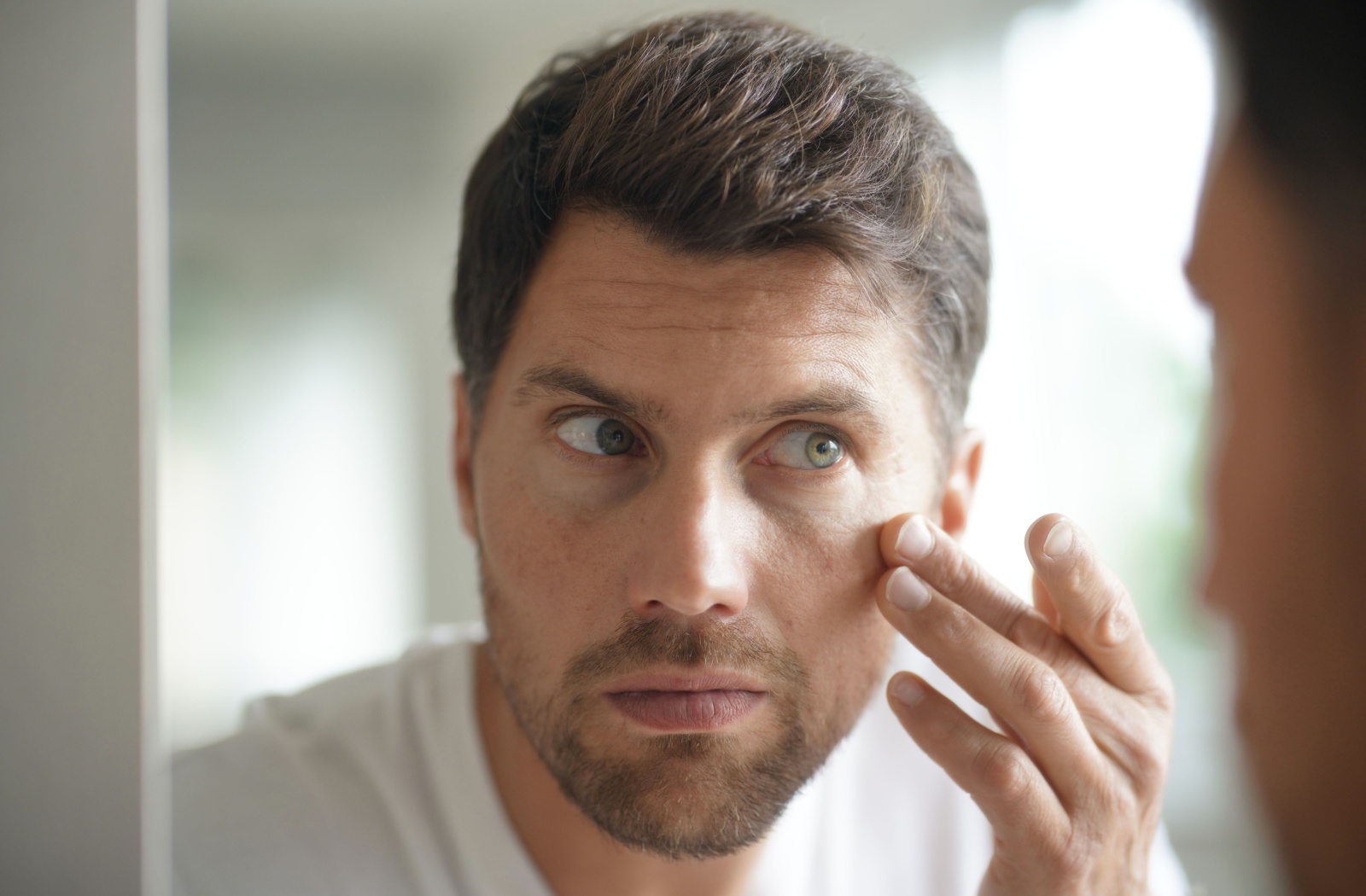 A person looking at their irritated eye in the mirror to find the cause of the irritation