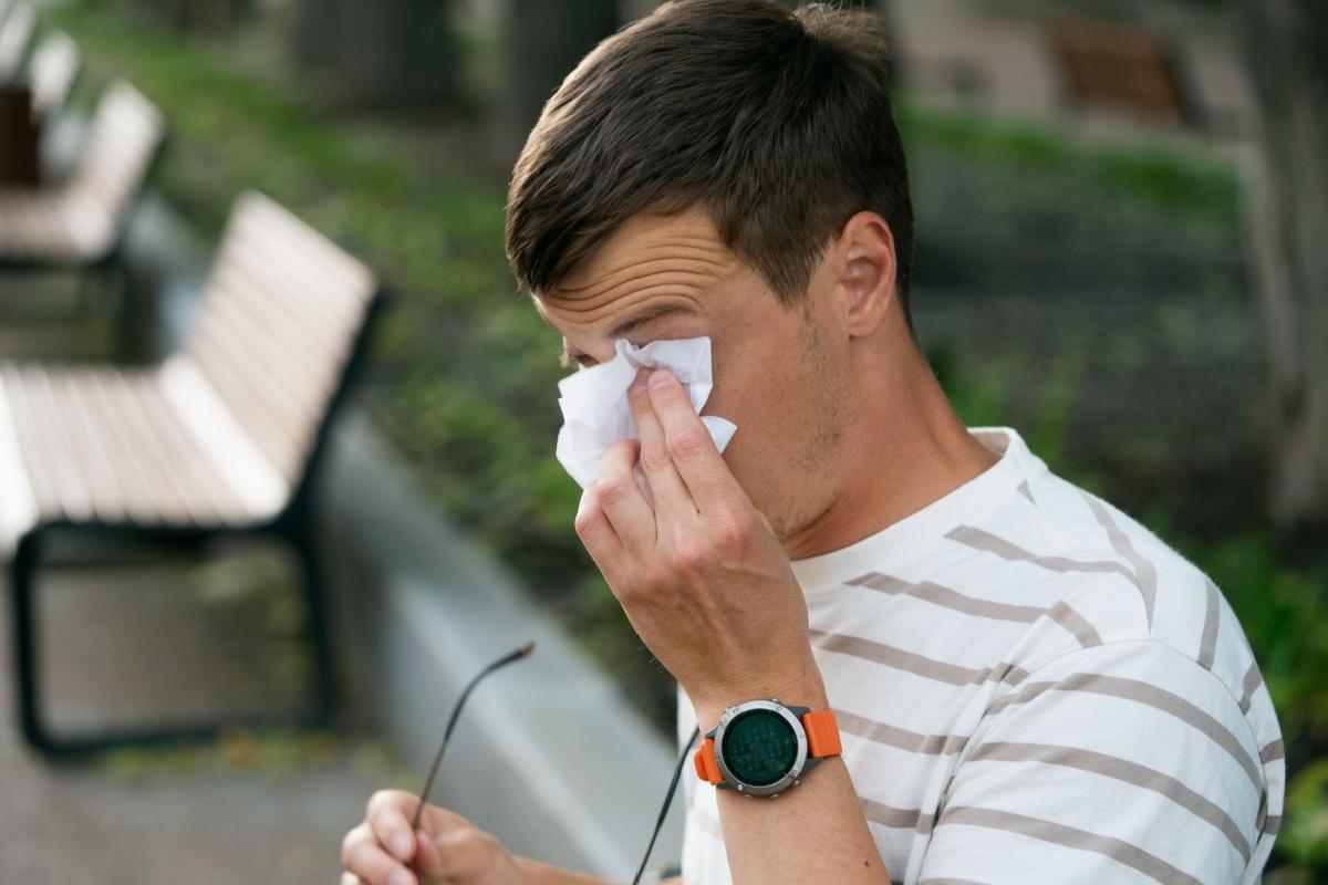 A person sitting on a park bench and using a tissue to rub their eye that is irritated from allergies