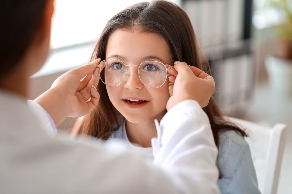 Eye care professional fitting a young girl with round eyeglasses during a pediatric eye exam in a bright optometry clinic.